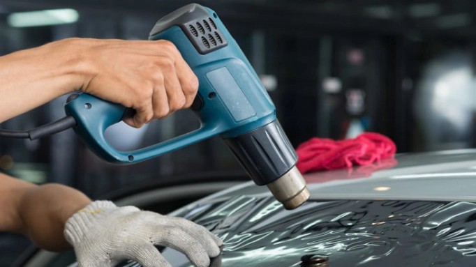 Mechanic using a cordless heat gun to apply vinyl wrap to a car panel in a garage setting